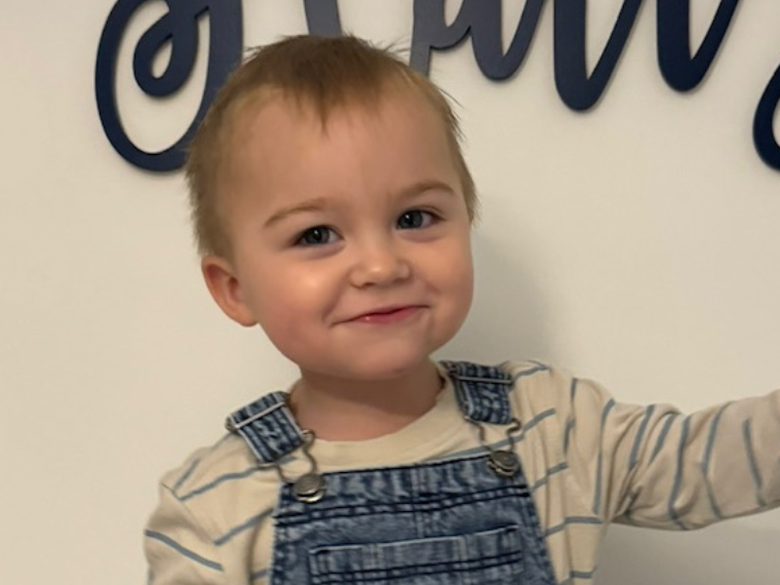 A toddler with blonde hair, wearing denim dungarees, smiles to camera
