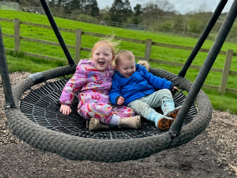A young girl puts her arm round her toddler brother, on a large swing in a park